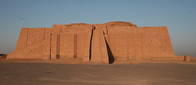 A large stepped ziggurat built of mudbrick rises from the desert landscape, featuring multiple terraced levels and a central staircase leading upward. The ancient structure shows weathered surfaces and strong geometric lines, representing the monumental architecture of early Mesopotamia.