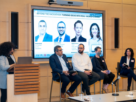 Panel discussion with four people seated, one speaking into a microphone. Background features a screen with "BEYOND THE HACKATHON"