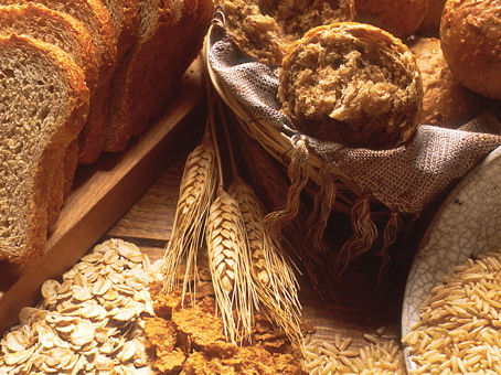 Golden wheat, flakes of oatmeal, and other grains are spilled on a table around loafs of sliced homemade bread.