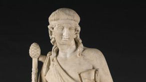 A statue of the God Dionysus as a young man, holding a chalice of wine in His left hand and a thyrsus staff in His right hand. He is wearing a sash of cloth around his upper torso, and is otherwise naked.