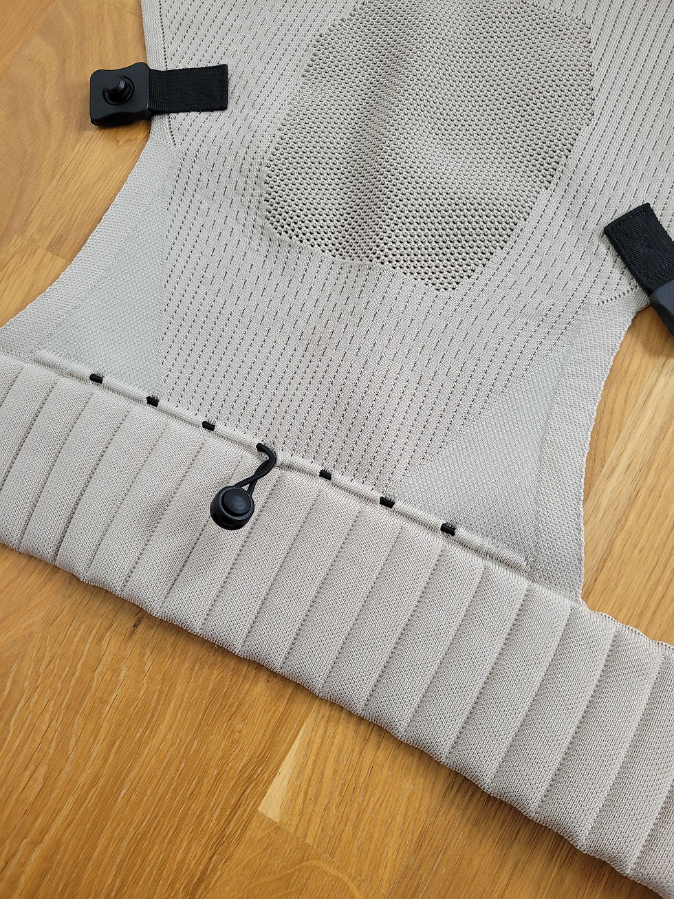 Beiger Strickbezug mit schwarzen Clips und Kordel auf Holzfußboden, nah aufgenommen. Testbericht Flybaby Babytrage