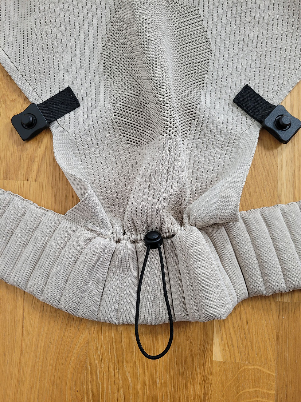 Nahaufnahme eines hellgrauen Strickbezugs mit schwarzen Clips und Kordel auf Holzboden. Testbericht Flybaby Babytrage