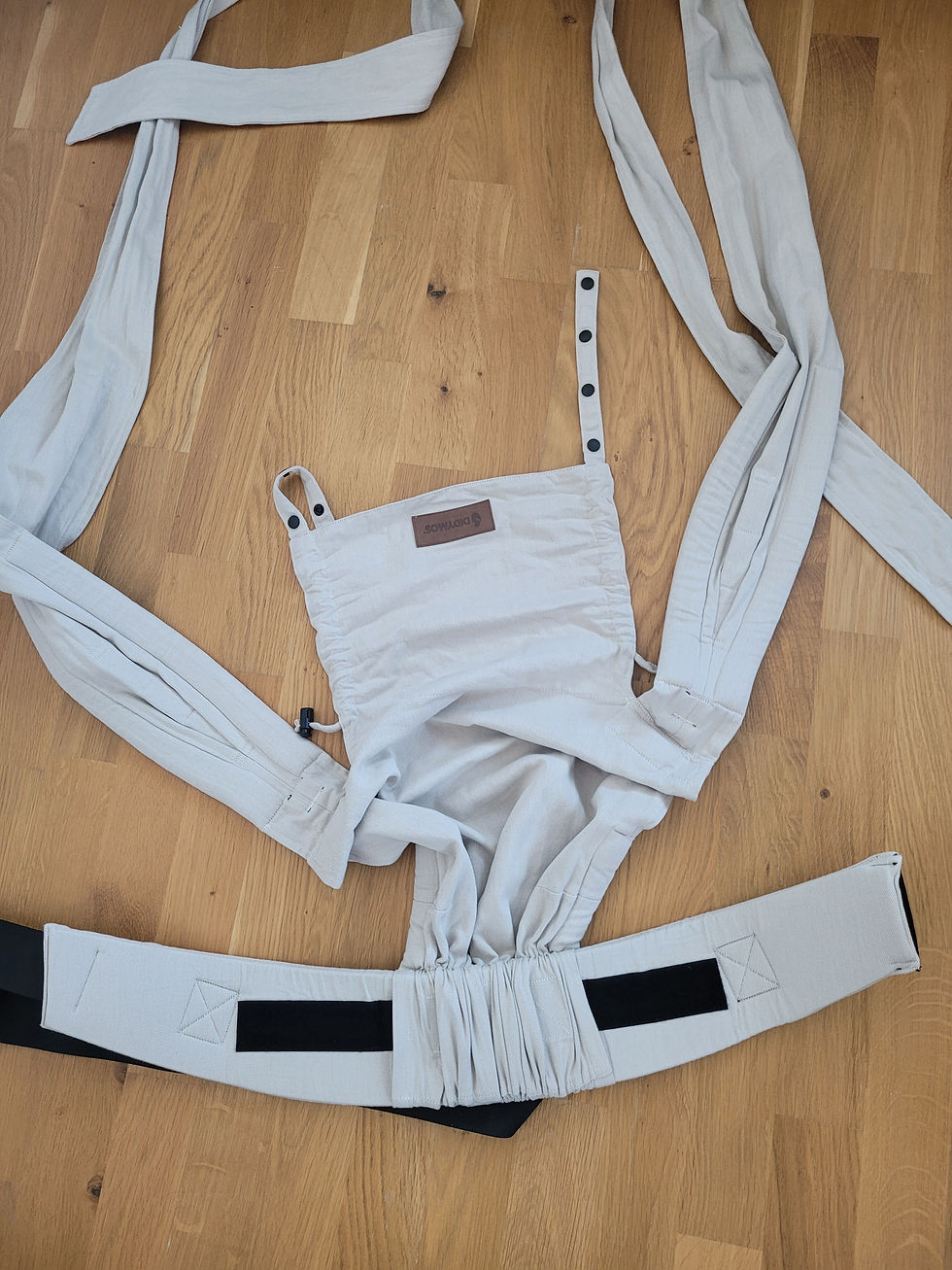 Weiße Babytrage mit schwarzem Gurt und Etikett auf Holzboden ausgebreitet. Didymos Didyflow Babytrage im Design Zimt