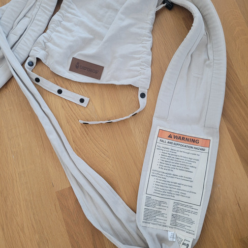 Didymos Didyflow Babytrage im Design Zimt. Die Trage liegt am Boden. Auf der Innenseite des Trägers ist ein Sicherheitslabel aufgenäht