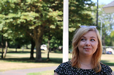 A white woman with a park in the background. Woman has mid-length blond hair. She has a pair of glasses on top of her head. She is wearing a black shirt with small floral pattern, and a minimal triangle necklace.