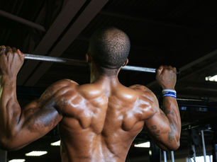 Muscular male athlete performing a pull-up on a bar in a gym, viewed from behind