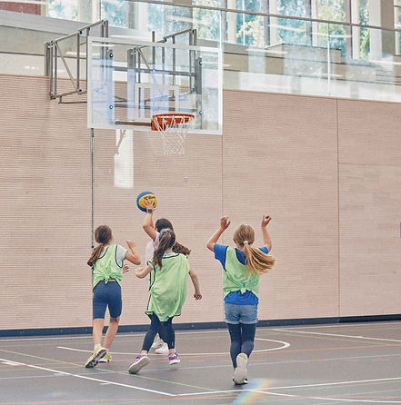 Sporthalle mit Basketballkorb, Ein Mädchen wirft den Ball zum Korb - drei Mädchen versuchen zu verteidigen