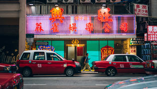 香港移民局 - 在城市环境中，红色出租车在一条熙熙攘攘的街道上排列在霓虹灯标志下。一个人在五颜六色的灯光背景下拿着手机走过。