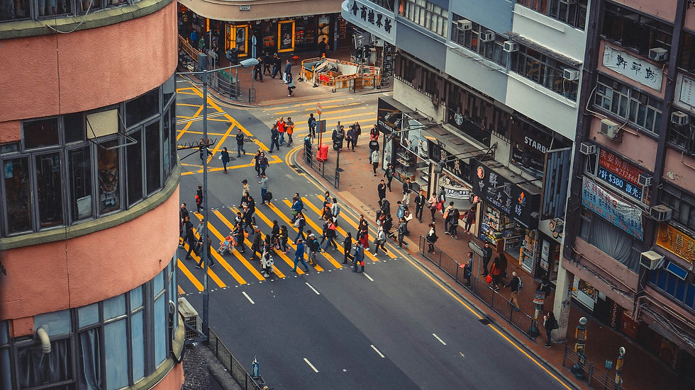 Hong Kong Immigration - Crowded crosswalk with people wearing jackets, surrounded by tall buildings and stores. Yellow stripes, urban hustle, overcast sky.