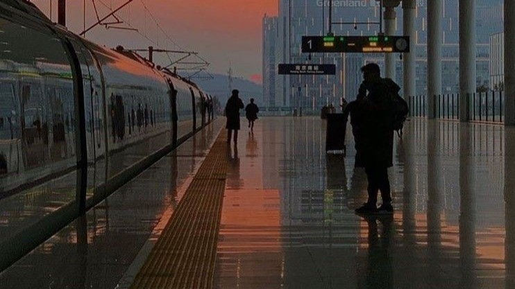 China Business Visa: Chinese Train station at sunset with a train on the left. Silhouetted people walk on the platform. Reflections on the floor, orange-pink sky.