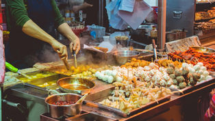 香港移民 - 供应商烹饪街头小吃，在热气腾腾的摊位上烧烤串，摊位上摆满了色彩缤纷的肉类和海鲜，营造出熙熙攘攘的市场氛围。