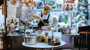 Hong Kong Work Visa: Chef cooking at a busy street food stall, surrounded by pots, utensils, and food displays. Menu signs and vibrant colors fill the scene.