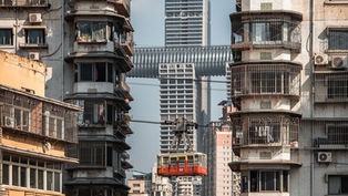 China Immigration. Chongqing: A red cable car moves between two old buildings with barred windows. A modern skyscraper with a skybridge looms in the background.