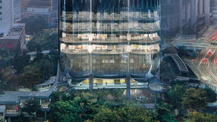 Hong Kong QMAS: The Henderson at dusk, with illuminated interiors showing people on each floor. Surrounding cityscape and roads are visible.