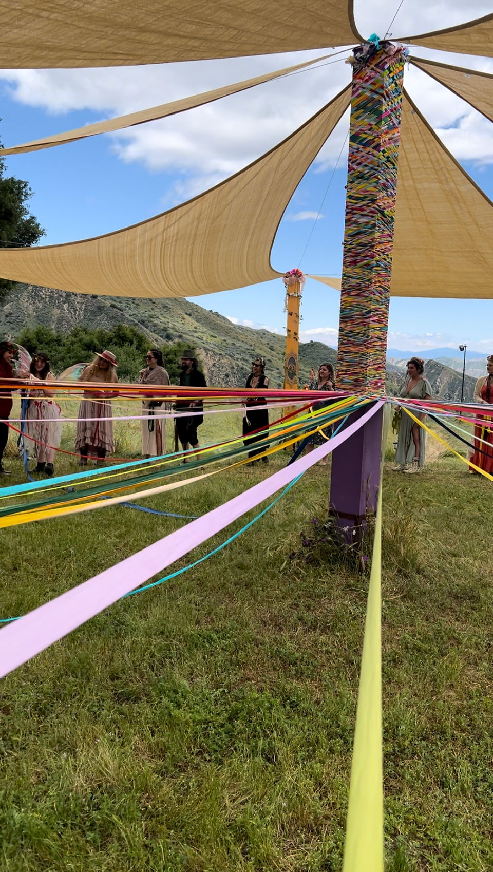 A beautiful wooden pole adorned with ribbons surrounded by people in a grassy field, dancing the may pole