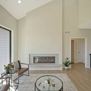 Bright modern living room with high vaulted ceiling, electric fireplace in white wall, leather chairs, glass coffee table, and large windows.