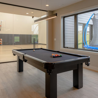 Game room with black pool table, rack of balls, glass wall showing indoor basketball court, and window view of trampoline outside.
