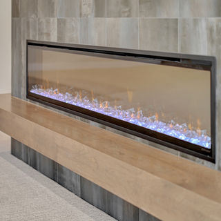 Modern linear fireplace with blue glass crystals, set in gray tiled wall, wooden ledge below, and light carpeted floor nearby.