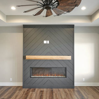 Modern living room with dark gray chevron-patterned fireplace, wooden mantel, long electric fire, rustic wood ceiling fan, and wood flooring.