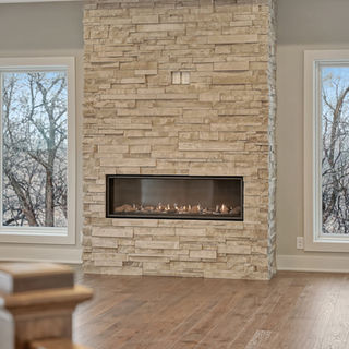 Modern room with stone fireplace, rectangular gas firebox, flanked by two large windows showing leafless trees, and hardwood floors.