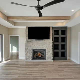 Spacious living room with stone fireplace, built-in black shelving, TV mounted above, wooden floor, beige walls, and ceiling fan with beams.