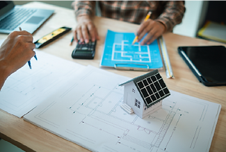 Two people working on a house design, with one person drawing on a blueprint, a solar-powered model house on the desk, and a calculator and laptop visible.