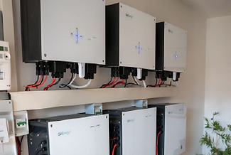 Several solar inverters mounted on a wall with wiring connections visible, next to an energy meter and a small plant.