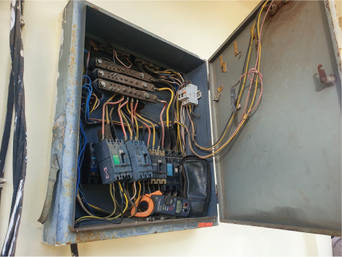 Open electrical panel showing exposed wires and switches with a multimeter inside. The metal box is weathered with rust spots.