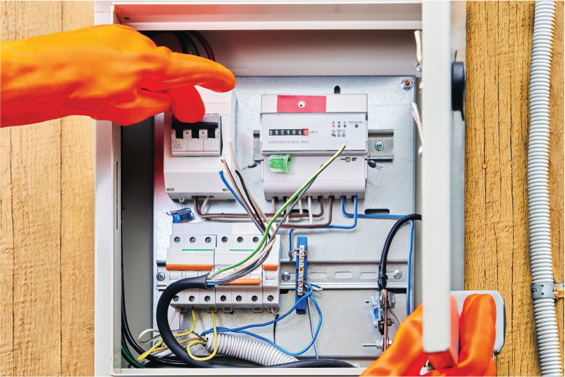 Hands in orange gloves adjust wires inside an electrical panel against a wooden wall. The panel features switches and a meter.