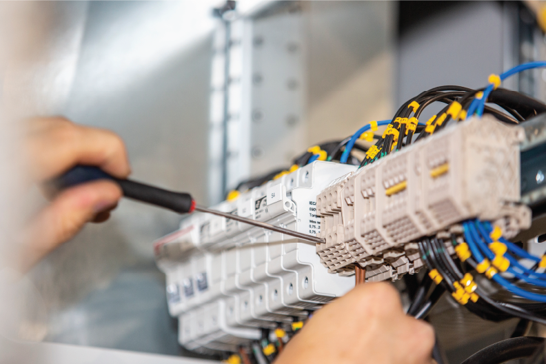 Hands use screwdriver on electrical panel with numerous wires and circuit breakers. Close-up of tools and components in technical setting.