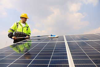 A worker in a yellow hard hat and high-visibility jacket cleaning a solar panel using a long-handled brush, with blue skies in the background.