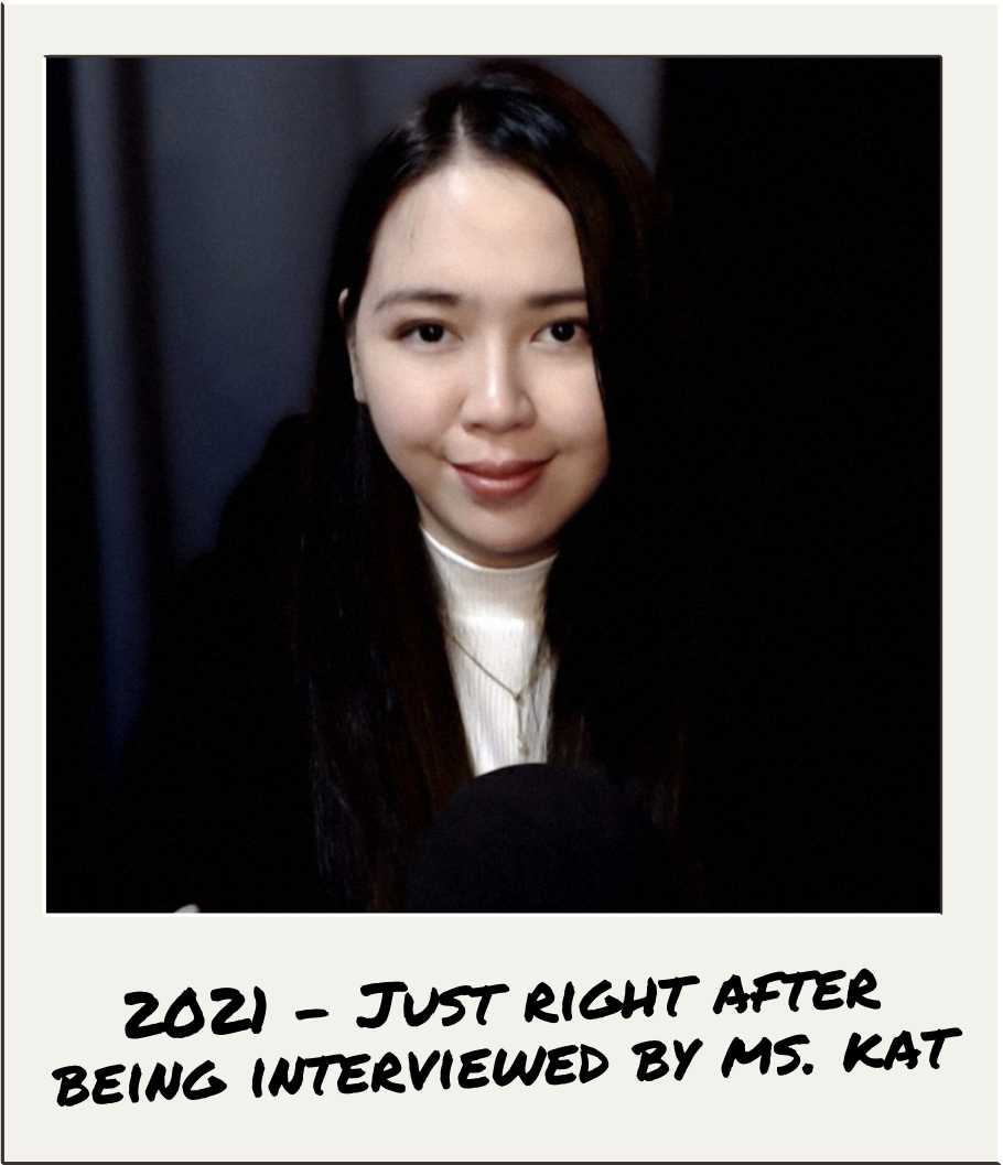 Smiling woman with long dark hair sits against a gray curtain backdrop, wearing a black jacket and white top. Text: "2021 - Just right after being interviewed by Ms. Kat."