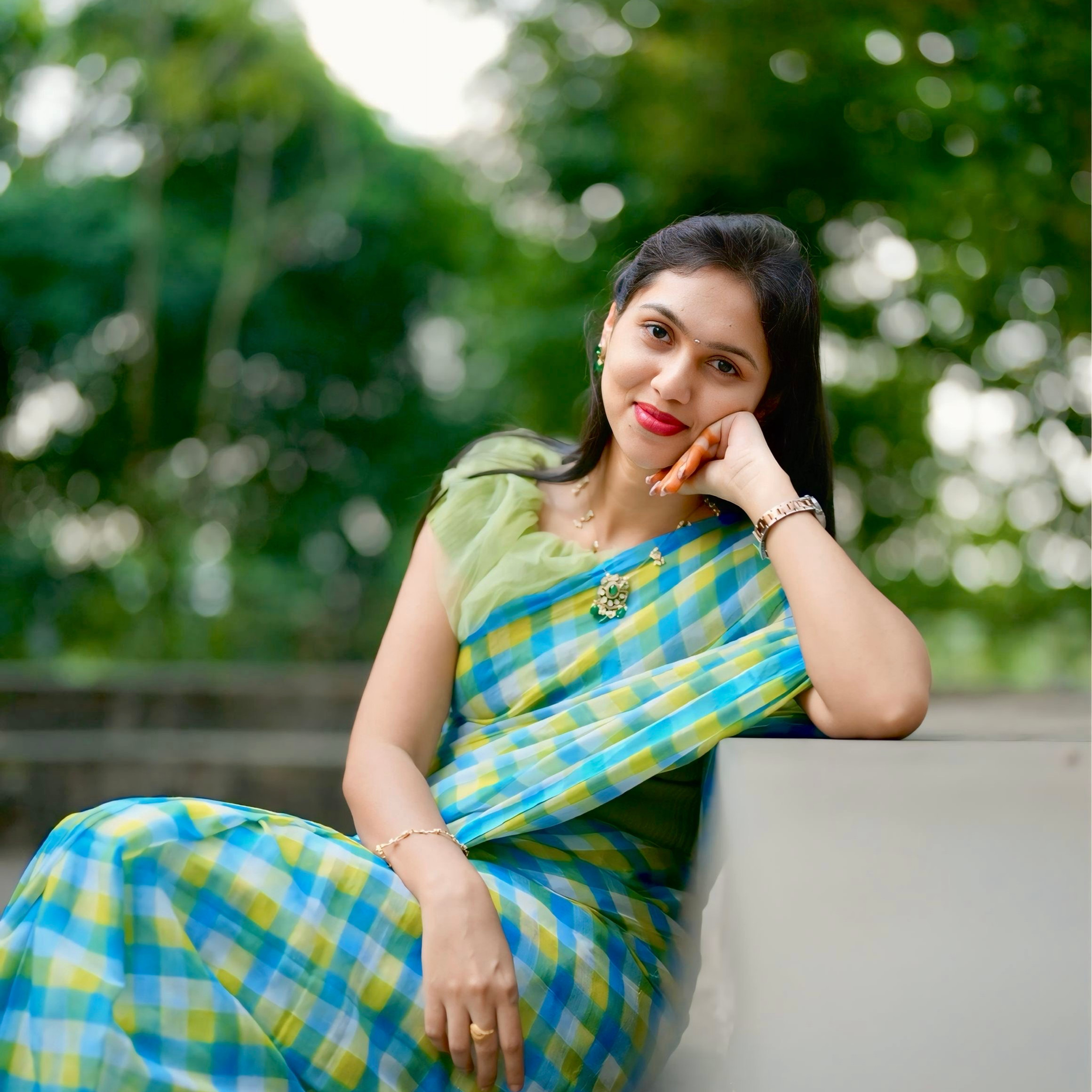 Smiling Sunitha Komarshetty in blue-green plaid sari sits on a concrete bench outdoors, resting her chin on her hand against a blurred green background.
