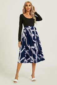 Woman in long-sleeved black top and blue patterned skirt stands against a plain background, wearing white sandals, looking confident.