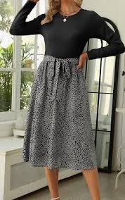 Woman in black top and patterned skirt standing in a bright room with plants and a mirror. Casual and stylish atmosphere.