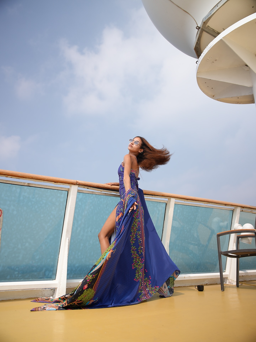 Nancy Tyagi in sunglasses poses on a ship deck, blue patterned dress flowing in the wind under a bright sky.