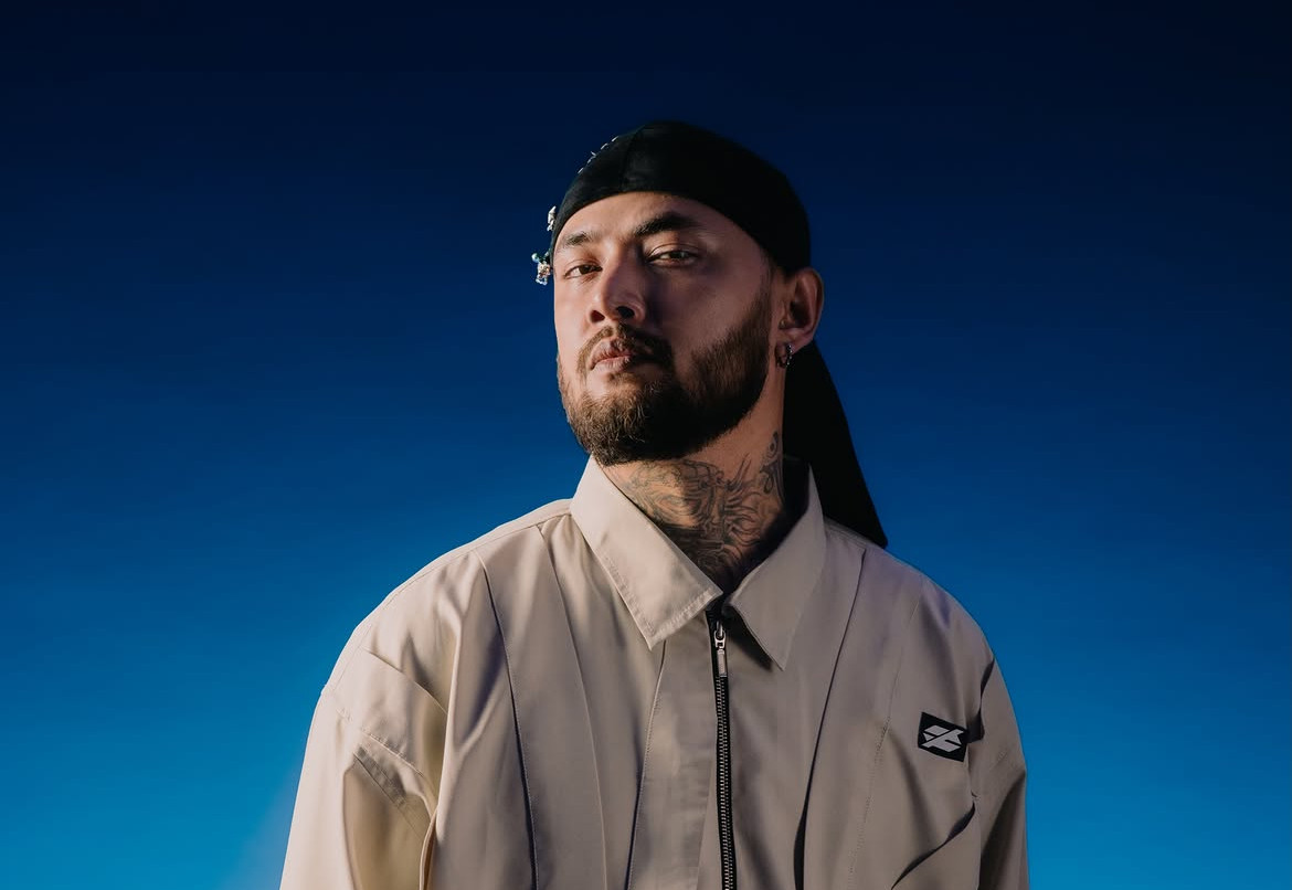 Portrait of a Raga in a beige tracksuit and black durag against a blue gradient background, with neck tattoos and a logo patch