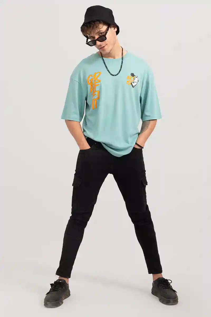Man in sunglasses and a black hat poses confidently. Wearing a teal shirt with orange graffiti, black pants, and sneakers. Gray background.