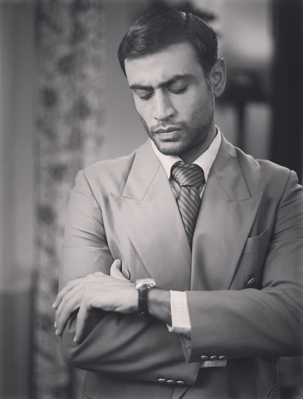 Black-and-white portrait of Armaan Khera in a suit, eyes closed, checking his wristwatch against a blurred indoor background.
