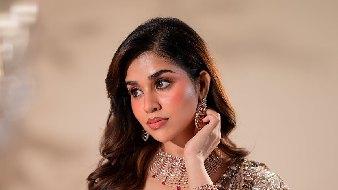 Portrait of Meenakshi Govindharajanin a sparkling pink-gold lehenga adjusting her earring against a soft beige studio backdrop.