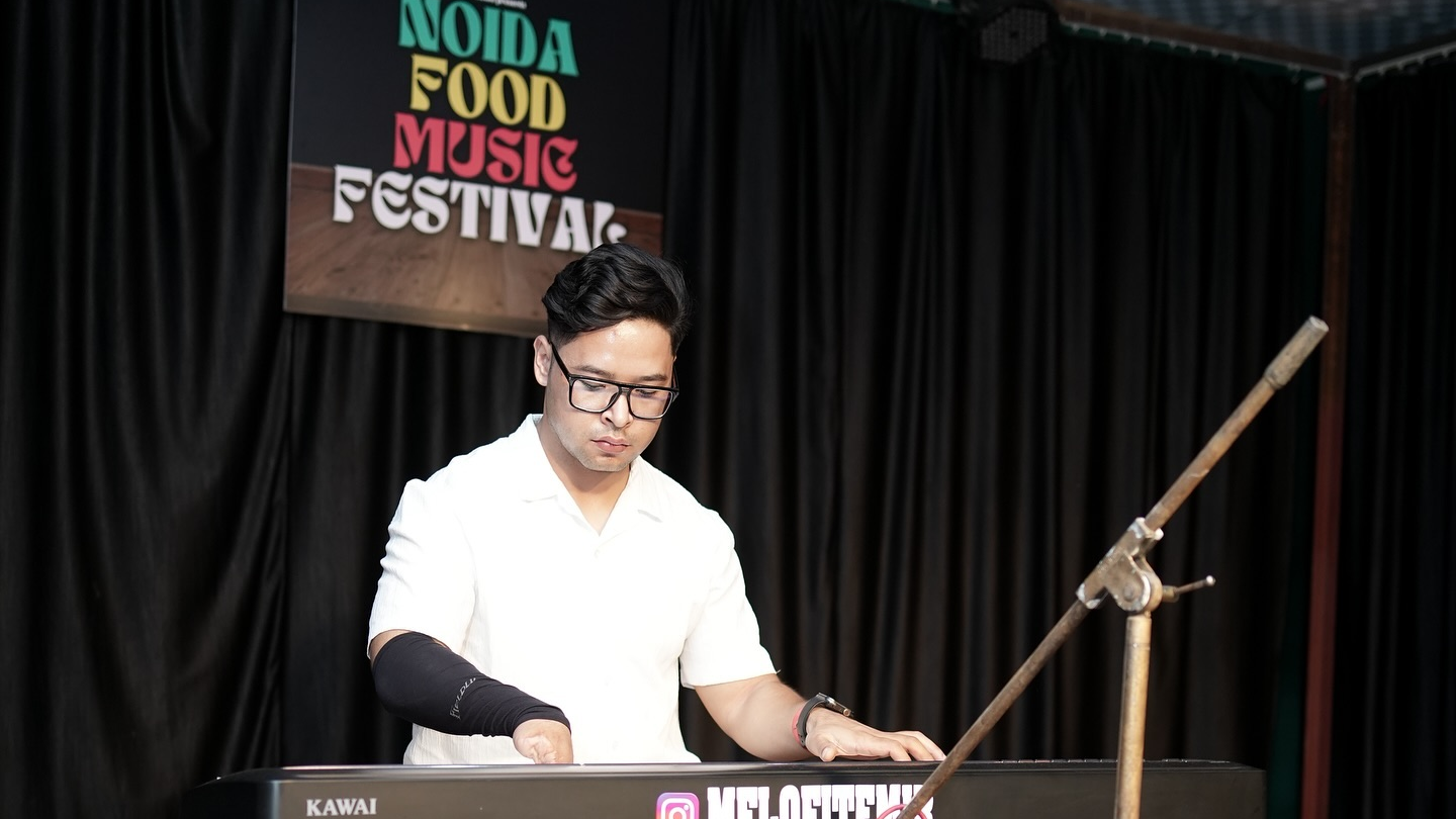 Melofit Emir in white shirt plays a Kawai keyboard onstage at Noida Food Music Festival, with black curtains and a focused mood.