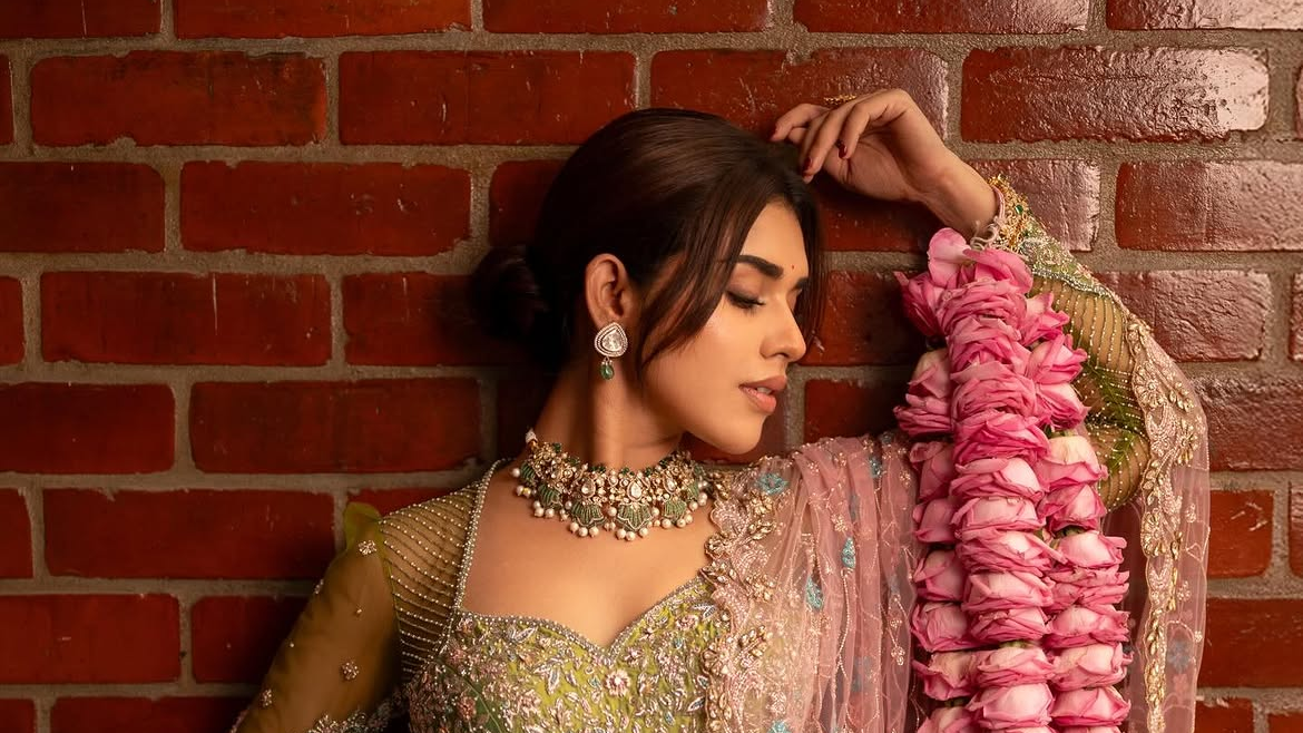 Meenakshi Govindharajan in ornate green lehenga with pink flower garlands poses against a red brick wall, eyes closed in a serene mood.