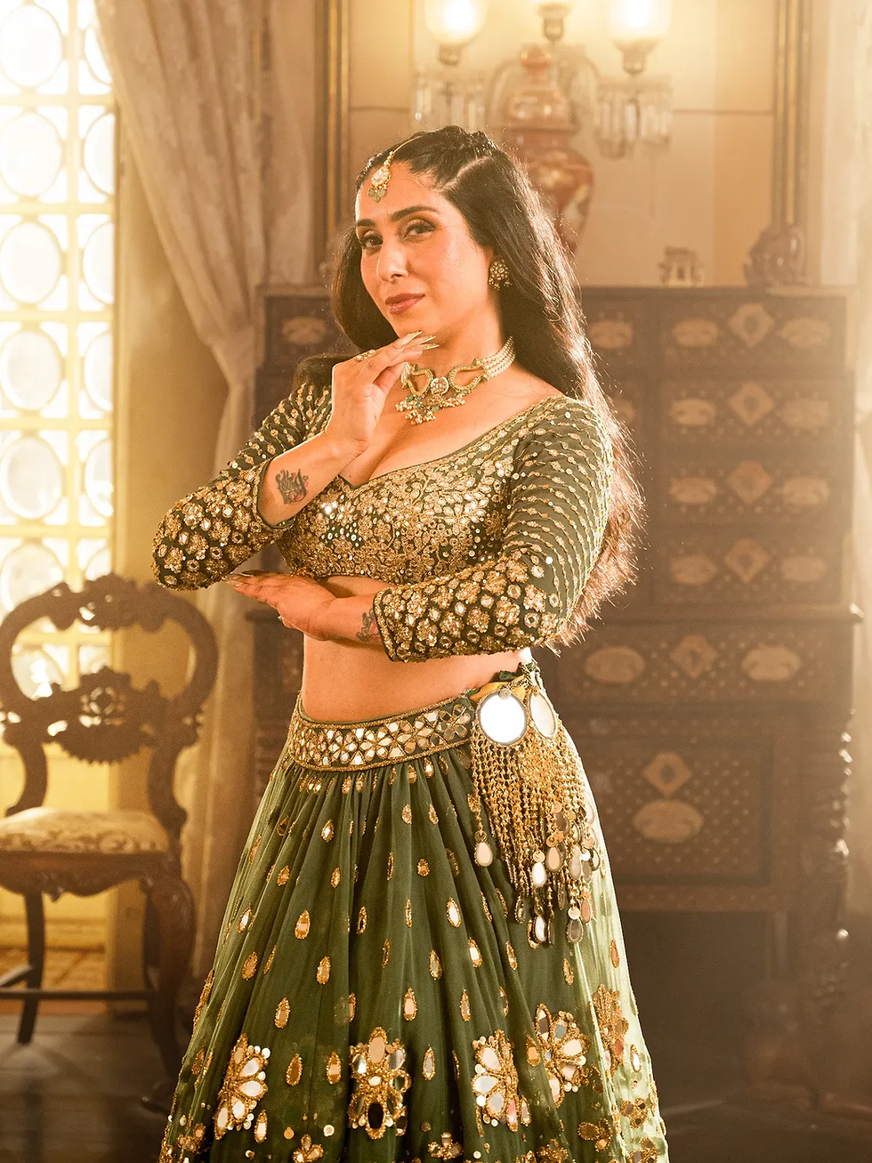 Neha Bhasin in ornate green lehenga and gold jewelry poses in a warm-lit room, hand under chin, with carved chair and drapes behind.