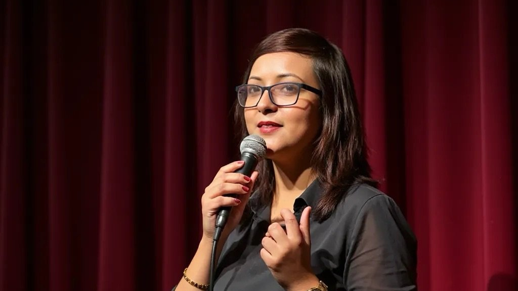 Banti Banerjee with glasses speaks into a microphone on stage before red curtains, wearing a black outfit.