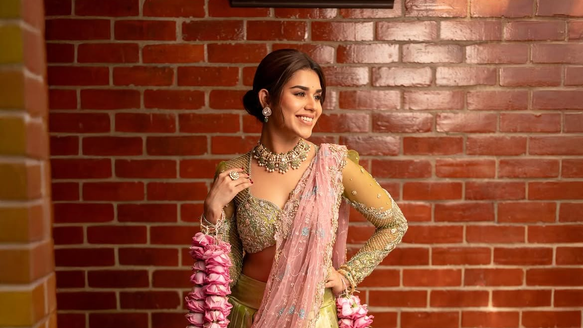 Smiling Meenakshi Govindharajan in a pink and green embroidered lehenga with floral garlands stands against a red brick wall.