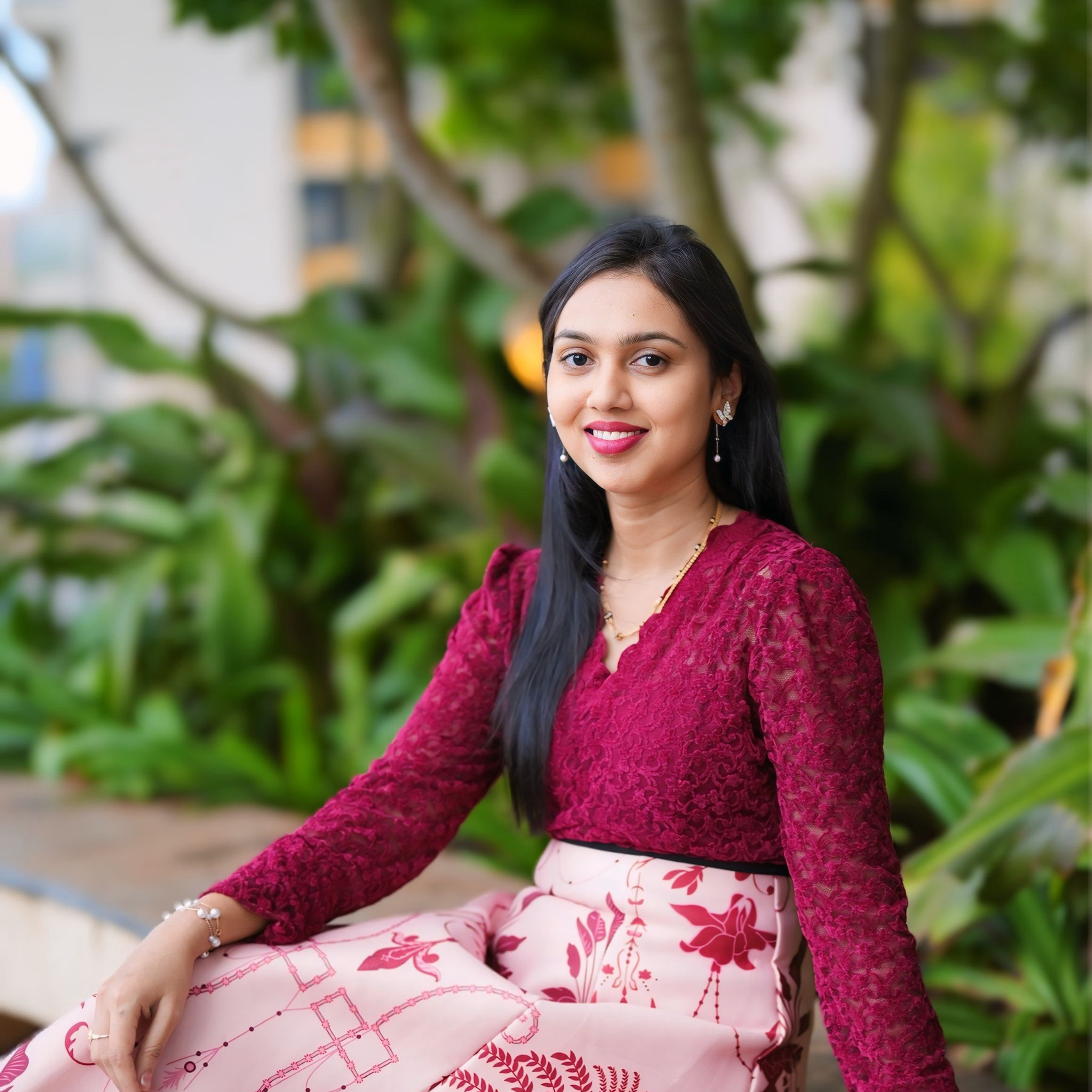 Smiling Sunitha Komarshetty in a magenta lace top and pink floral skirt sits on a bench in a lush green outdoor setting.