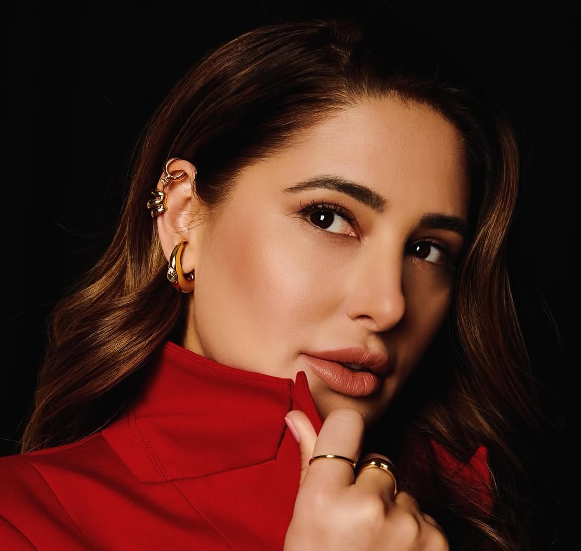 Portrait of Nargis Fakhri in a red jacket with gold earrings, posing against a black background with a calm, confident expression