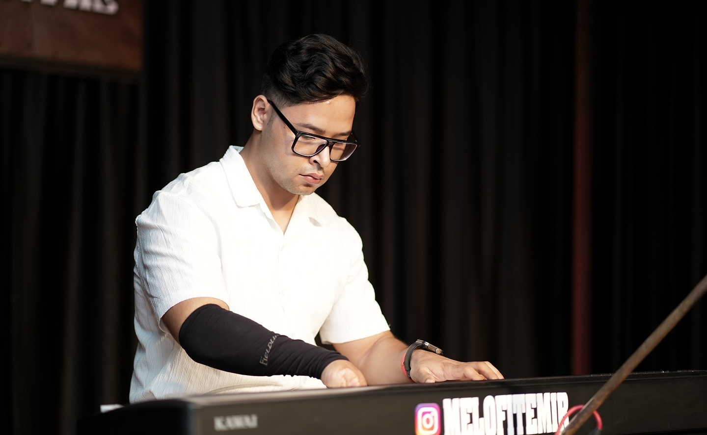 Melofit Emir in glasses and white shirt plays keyboard on a dark stage, focused, with an Instagram handle visible on the instrument.