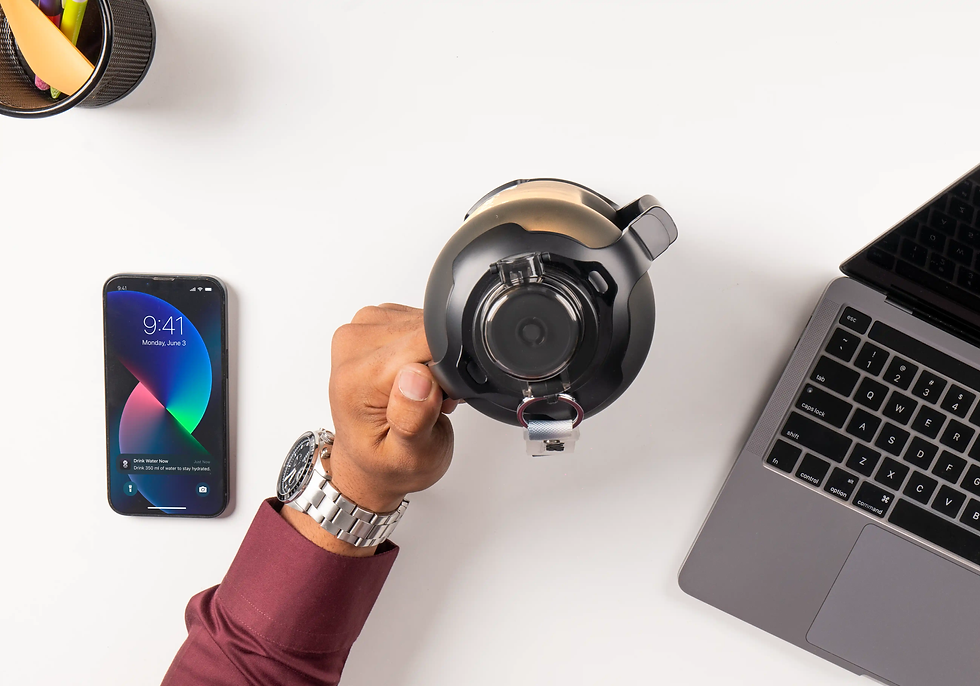 Hand holding a black kettle on a white desk with a laptop and smartphone showing 9:41. Alert reads: "Drink Water Now." Office setting.