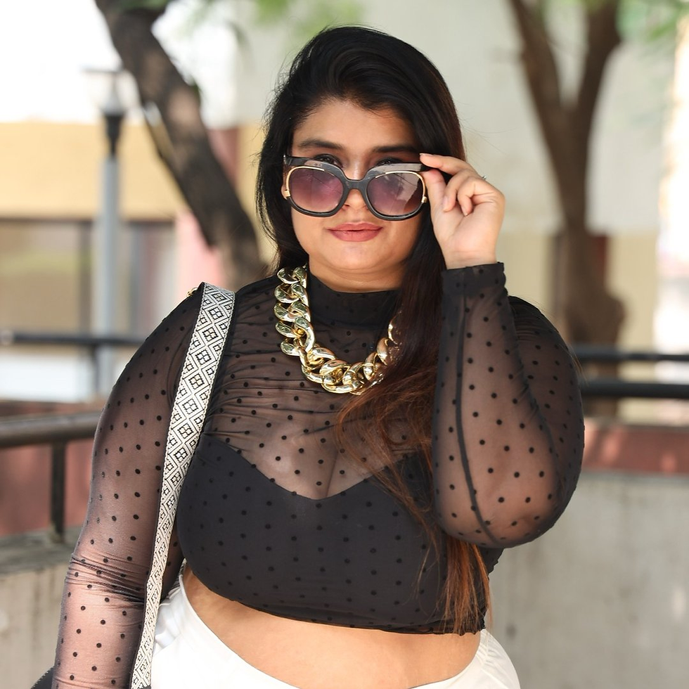 Darshana Bulchandani in oversized sunglasses adjusts them outdoors, wearing a black sheer polka-dot top and gold chain necklace.